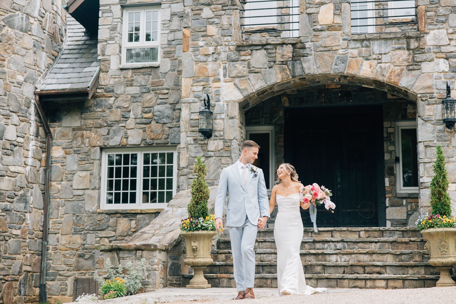 Bride and groom walk hand-in-hand, smiling in front of a stone castle-style venue.